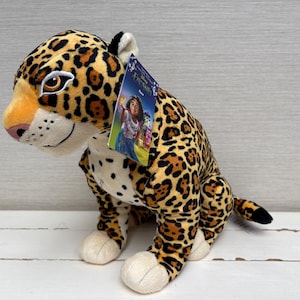 May include: A plush jaguar toy with a yellow and black spotted coat, white underbelly, and brown eyes. A rectangular Disney Encanto tag featuring a character illustration is attached to the side of the head. The toy is in a seated position.