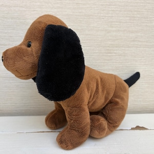 May include: A plush toy dog with soft brown fur and contrasting black ears. The stuffed animal is in a seated position, featuring a small black nose, dark round eyes, and a short black tail extending from the back of its brown body.