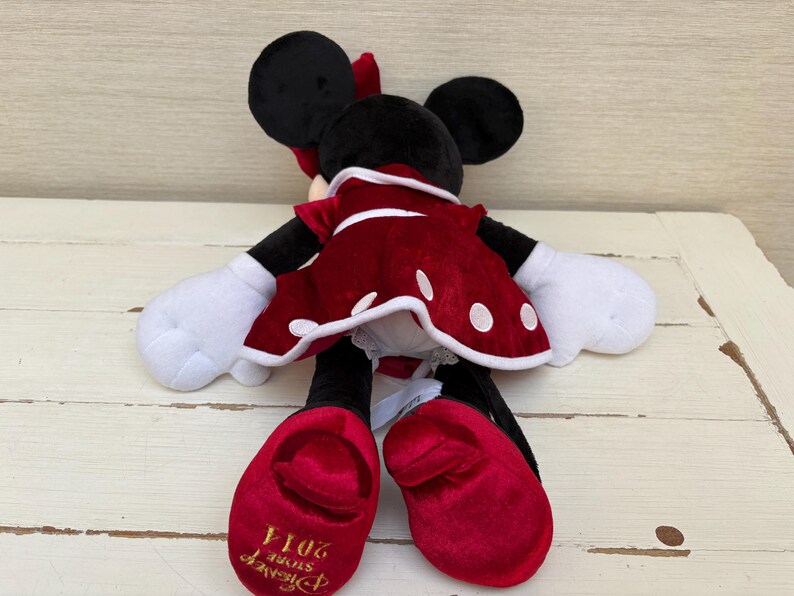 May include: A plush Minnie Mouse doll, with black ears, a red bow, and a red dress with white polka dots. The doll has white gloves and red shoes. The Disney Store 2014 logo is visible on the shoe.