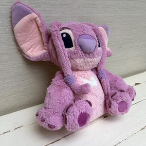 May include: A plush toy of a character with large ears, purple fur, and pink accents. The toy has a light purple nose, dark eyes, and long, braided hair. The character is sitting on a white surface.