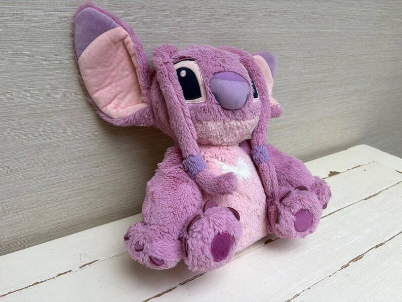 May include: A plush toy of a character with large ears, purple fur, and pink accents. The toy has a light purple nose, dark eyes, and long, braided hair. The character is sitting on a white surface.