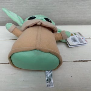 Peut inclure: Une peluche d'un personnage de la franchise Star Wars. Le jouet a de grands yeux noirs, une peau verte et de grandes oreilles. Il porte un manteau beige et a un corps vert. Une &eacute;tiquette est attach&eacute;e au jouet.
