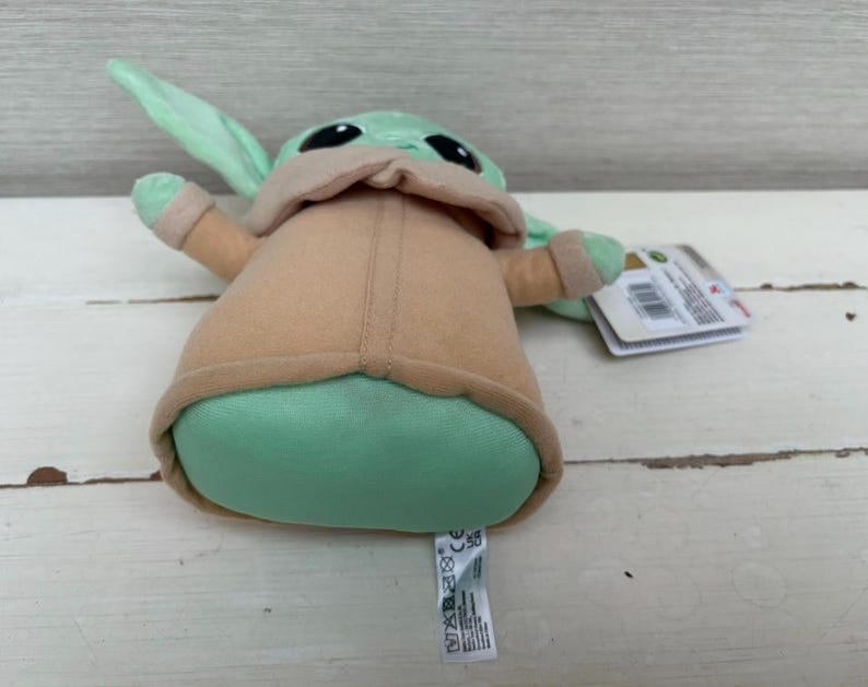 Peut inclure: Une peluche d'un personnage de la franchise Star Wars. Le jouet a de grands yeux noirs, une peau verte et de grandes oreilles. Il porte un manteau beige et a un corps vert. Une &eacute;tiquette est attach&eacute;e au jouet.