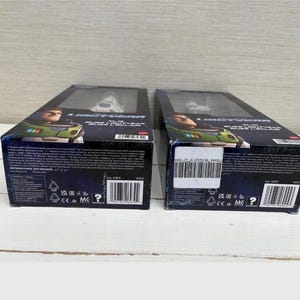 May include: Two boxed Buzz Lightyear action figures. The boxes are black with images of Buzz Lightyear and the film title. The figures are visible through a clear plastic window. The boxes are on a light wooden surface.