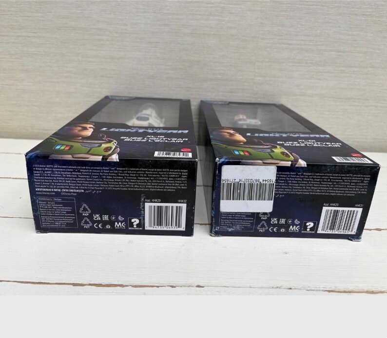 May include: Two boxed Buzz Lightyear action figures. The boxes are black with images of Buzz Lightyear and the film title. The figures are visible through a clear plastic window. The boxes are on a light wooden surface.