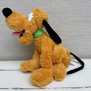 Puede incluir: Un peluche de Pluto, un personaje de dibujos animados. El juguete es principalmente naranja con orejas y cola negras, una nariz negra y una lengua rosa. Tiene un collar verde con las letras "TO".