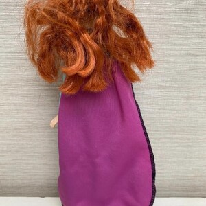 May include: A doll with vibrant red curly hair and a purple cape with black edging. The doll's back is visible, showing the cape and hair in detail. The doll's arm is visible.