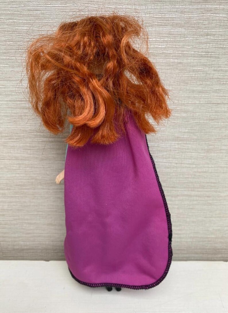May include: A doll with vibrant red curly hair and a purple cape with black edging. The doll's back is visible, showing the cape and hair in detail. The doll's arm is visible.