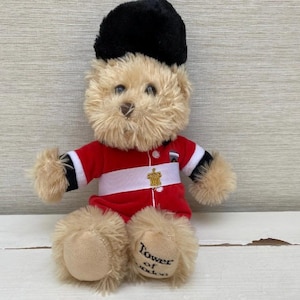 May include: A plush teddy bear wearing a red and white uniform with a black hat. The bear has tan fur and the words "Tower of London" are printed on one foot. A gold emblem is on the uniform.