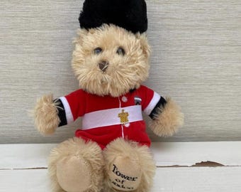 Könnte beinhalten: Ein Teddybär in roter und weißer Uniform mit schwarzem Hut. Der Bär hat hellbraunes Fell und die Aufschrift "Tower of London" auf einem Fuß. Ein goldenes Emblem befindet sich auf der Uniform.