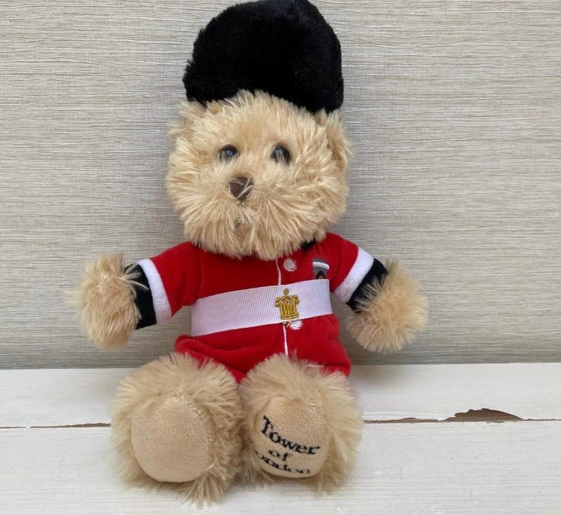 May include: A plush teddy bear wearing a red and white uniform with a black hat. The bear has tan fur and the words "Tower of London" are printed on one foot. A gold emblem is on the uniform.