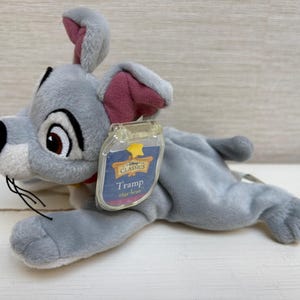 May include: A soft toy of Tramp from Disney's "Lady and the Tramp". The grey dog has a black nose, brown eyes, and pink inner ears. A clear tag with the Disney Classics logo and the name "Tramp" is attached.