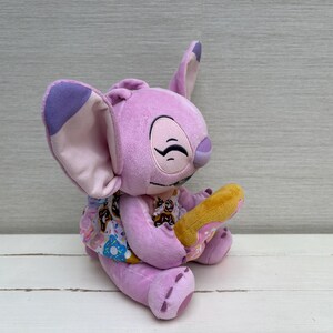 Puede incluir: Un peluche de color lavanda con orejas grandes y una cara sonriente, sosteniendo un donut amarillo. El personaje lleva un vestido estampado con im&aacute;genes de donuts. El juguete est&aacute; sobre una superficie blanca.