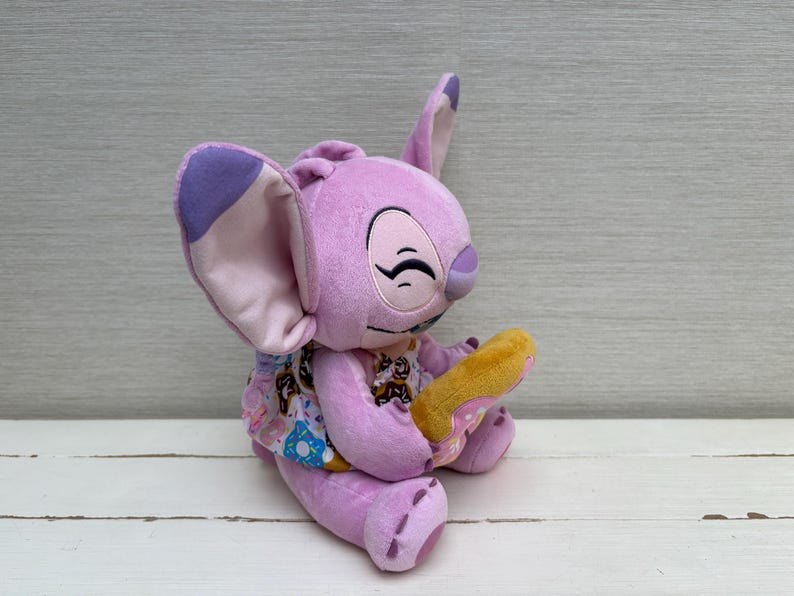 Puede incluir: Un peluche de color lavanda con orejas grandes y una cara sonriente, sosteniendo un donut amarillo. El personaje lleva un vestido estampado con im&aacute;genes de donuts. El juguete est&aacute; sobre una superficie blanca.