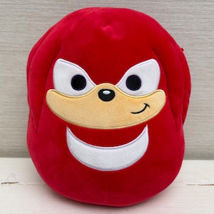 May include: A red plush character head featuring white eyes, a tan muzzle, and a white crescent-shaped mouth. The character has a black nose and a smiling expression. This is likely a toy or decorative item.