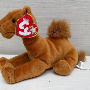 May include: A brown plush camel toy with a red heart-shaped tag. The tag has the text "Ty" and "2000" on it. The camel has a fluffy hump and tail. The toy is sitting on a light-coloured surface.