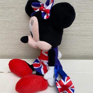 Puede incluir: Un peluche de Minnie Mouse vestida con un atuendo con el tema de la Union Jack. La mu&ntilde;eca tiene orejas negras, un lazo rojo y blanco y un vestido con la bandera brit&aacute;nica. Tiene zapatos rojos y guantes blancos.