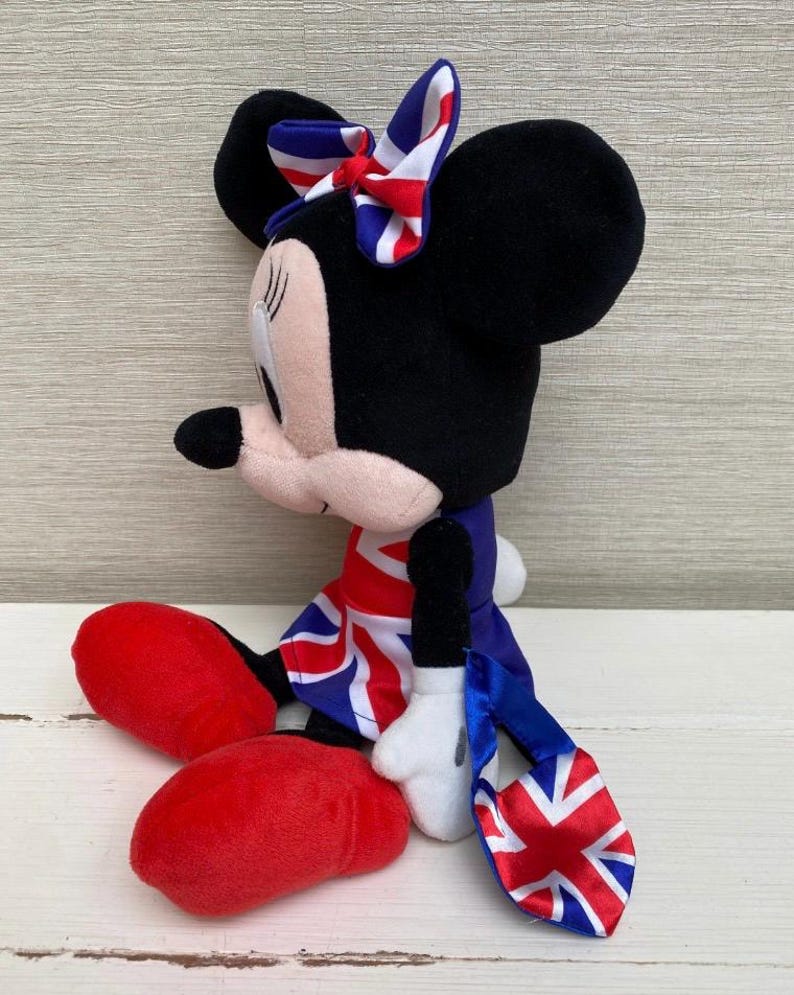 Puede incluir: Un peluche de Minnie Mouse vestida con un atuendo con el tema de la Union Jack. La mu&ntilde;eca tiene orejas negras, un lazo rojo y blanco y un vestido con la bandera brit&aacute;nica. Tiene zapatos rojos y guantes blancos.
