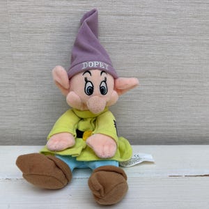 以下が含まれることがあります： ドーピーのキャラクターのぬいぐるみ。白糸で「DOPEY」と刺繍された薄紫色のとんがり帽子をかぶっています。長袖の黄色いチュニックに黒いベルトと金色のバックル、水色のズボン、茶色の靴を身につけています。