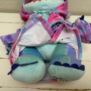 May include: A plush toy in shades of blue, purple, and pink, with a tie-dye pattern. The toy has large feet, claws, and a wide mouth. The toy is a character from Disney's Lilo & Stitch.