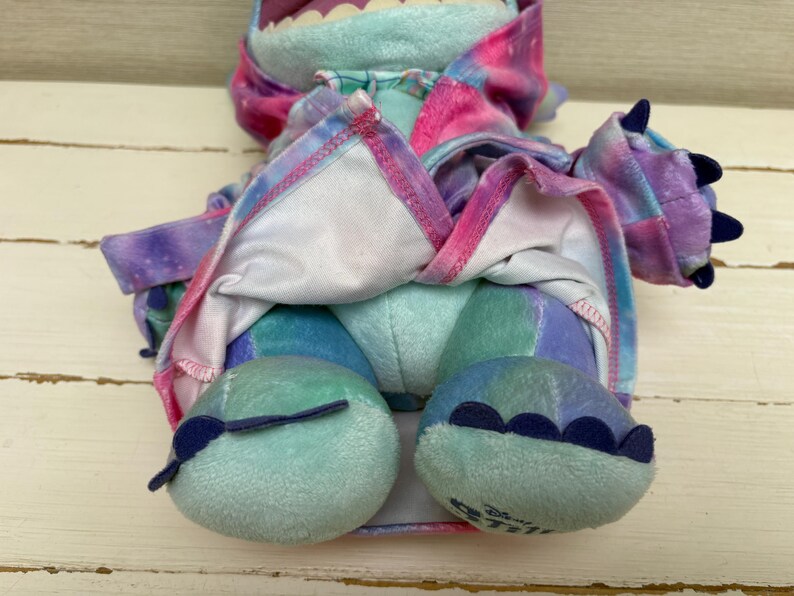 May include: A plush toy in shades of blue, purple, and pink, with a tie-dye pattern. The toy has large feet, claws, and a wide mouth. The toy is a character from Disney's Lilo & Stitch.