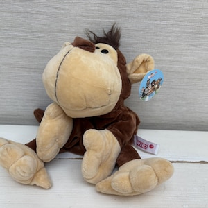 May include: A plush monkey toy with dark brown fur on its body and light beige fur on its face, hands, and feet. The monkey has a tuft of dark brown hair on its head, black eyes, and a round blue tag attached to its ear with the text NICI.