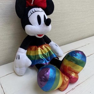 May include: A plush Minnie Mouse doll with a black body, white gloves, and a red bow. The doll features a rainbow-coloured skirt and rainbow-striped shoes, creating a vibrant and colourful design. The doll is sitting on a white surface.