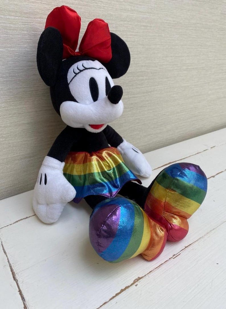 May include: A plush Minnie Mouse doll with a black body, white gloves, and a red bow. The doll features a rainbow-coloured skirt and rainbow-striped shoes, creating a vibrant and colourful design. The doll is sitting on a white surface.
