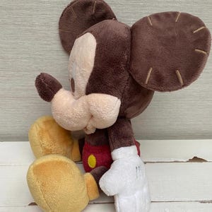 May include: A plush Mickey Mouse toy in a seated position. The toy has a brown head and ears, a tan face, and a red and yellow outfit. The hands are white, and the feet are yellow.
