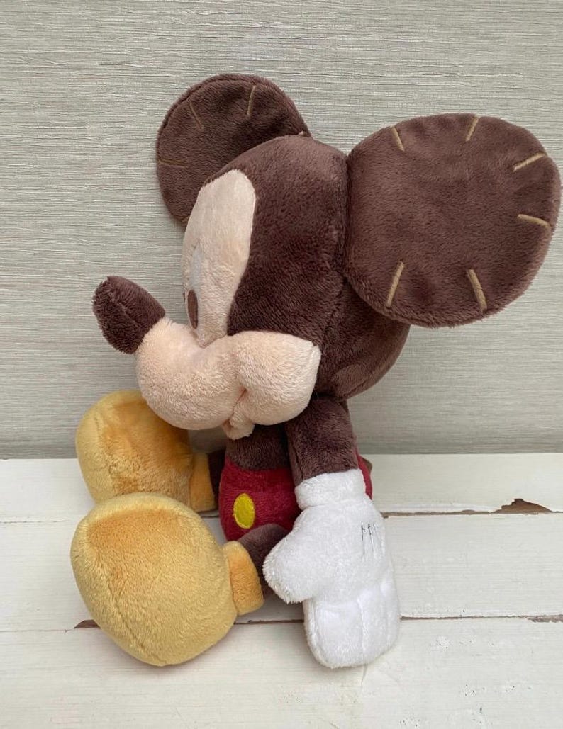 May include: A plush Mickey Mouse toy in a seated position. The toy has a brown head and ears, a tan face, and a red and yellow outfit. The hands are white, and the feet are yellow.