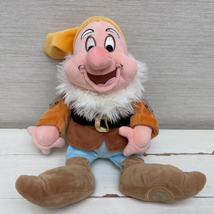 Könnte beinhalten: Ein Plüschtier der Figur Dopey aus Schneewittchen mit gelber Mütze, brauner Tunika mit schwarzem Gürtel und goldener Schnalle, hellblauer Hose und großen braunen Schuhen. Die Figur hat ein rosafarbenes Gesicht und einen weißen Kunstfellbart.