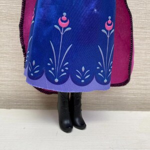 May include: A doll wearing a blue dress with a floral pattern and a purple hem. The dress has a pink and white floral design. The doll is wearing black boots and a magenta cape.