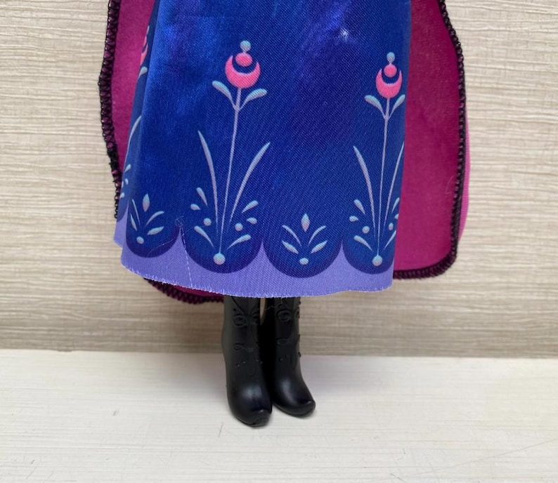 May include: A doll wearing a blue dress with a floral pattern and a purple hem. The dress has a pink and white floral design. The doll is wearing black boots and a magenta cape.