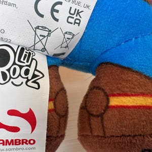 May include: Close-up of a plush toy with a blue upper body and brown legs. The legs have yellow and red stripes. A white tag displays the "Lil Bodz" logo and the Sambro logo. The toy is made in China.
