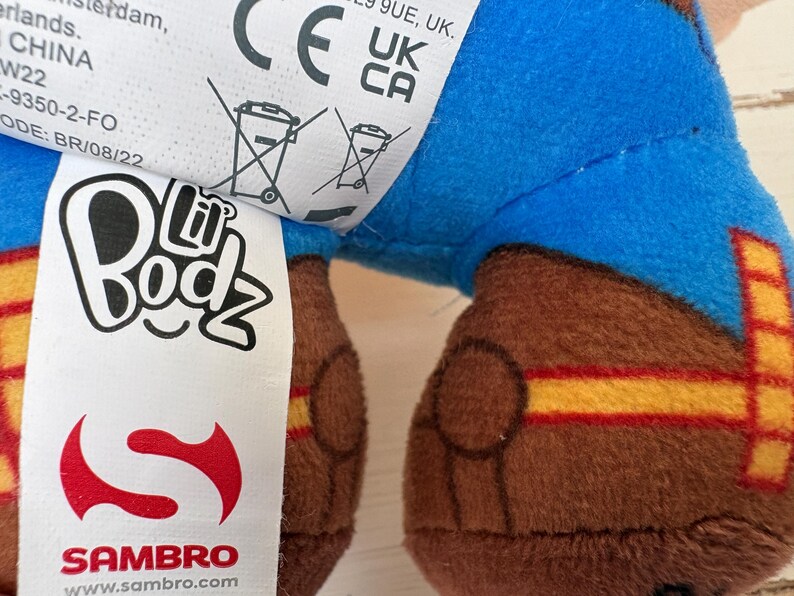 May include: Close-up of a plush toy with a blue upper body and brown legs. The legs have yellow and red stripes. A white tag displays the "Lil Bodz" logo and the Sambro logo. The toy is made in China.