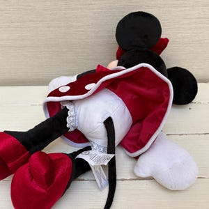 May include: A plush Minnie Mouse doll, lying on its side. The doll features a black head with large ears, a red bow, and a white and red polka-dot dress. The doll's large red shoes and white gloves are also visible.