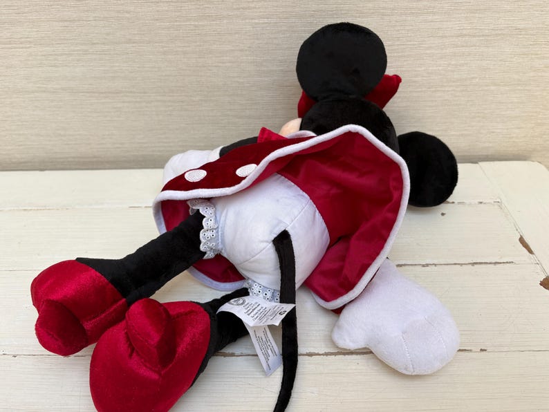 May include: A plush Minnie Mouse doll, lying on its side. The doll features a black head with large ears, a red bow, and a white and red polka-dot dress. The doll's large red shoes and white gloves are also visible.