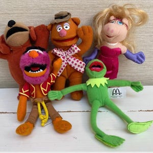 May include: A selection of Muppet Show plush toys, including Fozzie Bear, Miss Piggy, Kermit the Frog, Animal, and a brown dog character. The toys are brightly coloured and feature detailed facial expressions.