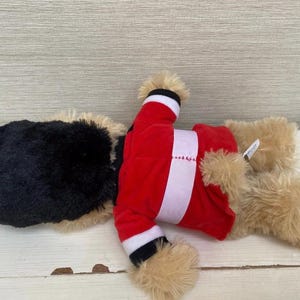 May include: A plush toy dog wearing a red Santa Claus outfit with white trim. The dog has a black head and tan fur on its paws and ears. The toy is lying on a white wooden surface.