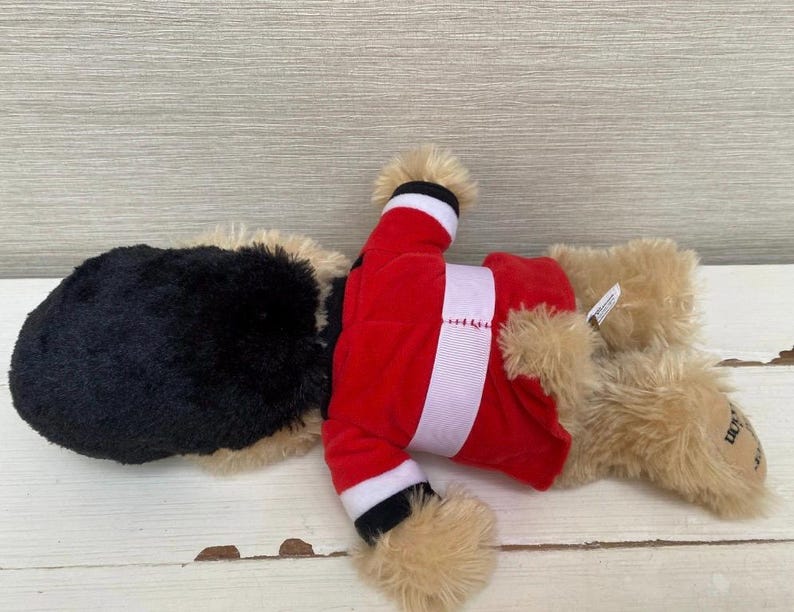 May include: A plush toy dog wearing a red Santa Claus outfit with white trim. The dog has a black head and tan fur on its paws and ears. The toy is lying on a white wooden surface.