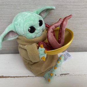 Pode incluir: Um brinquedo de pel&uacute;cia de um personagem de pele verde com orelhas grandes, vestindo uma t&uacute;nica marrom. O personagem segura uma tigela dourada com tent&aacute;culos vermelhos. O brinquedo tem olhos pretos grandes e pequenas garras amarelas.