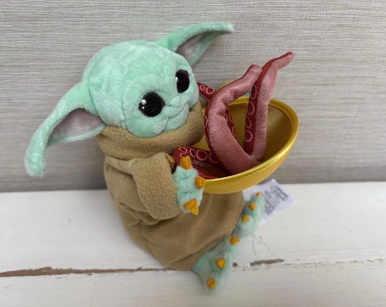 Pode incluir: Um brinquedo de pel&uacute;cia de um personagem de pele verde com orelhas grandes, vestindo uma t&uacute;nica marrom. O personagem segura uma tigela dourada com tent&aacute;culos vermelhos. O brinquedo tem olhos pretos grandes e pequenas garras amarelas.