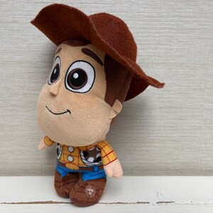 May include: A plush toy of a cartoon cowboy character, wearing a brown hat, yellow shirt with red details, blue pants, and brown boots. The toy has a smiling face and large eyes. The toy is a children's collectible.