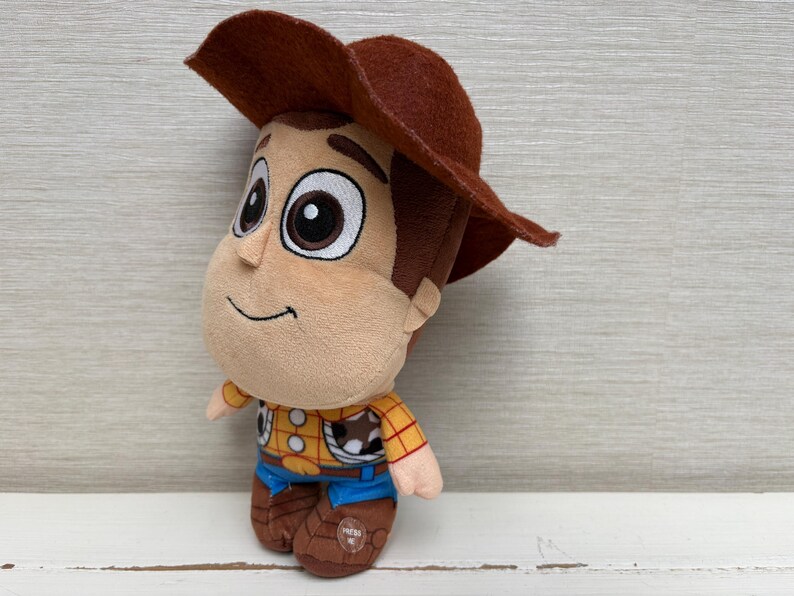 May include: A plush toy of a cartoon cowboy character, wearing a brown hat, yellow shirt with red details, blue pants, and brown boots. The toy has a smiling face and large eyes. The toy is a children's collectible.
