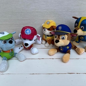 Può includere: Cinque pupazzi di cani dei cartoni animati, ognuno con uniformi e cappelli unici. I cani sono di vari colori, tra cui blu, rosso, giallo e marrone. I giocattoli sono disposti su una superficie di legno bianco.