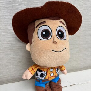 May include: A plush toy of a cartoon character with a brown cowboy hat. The toy has a tan face, brown hair, and large black eyes. It wears a yellow plaid shirt, blue pants, and brown boots. The character has a friendly smile.