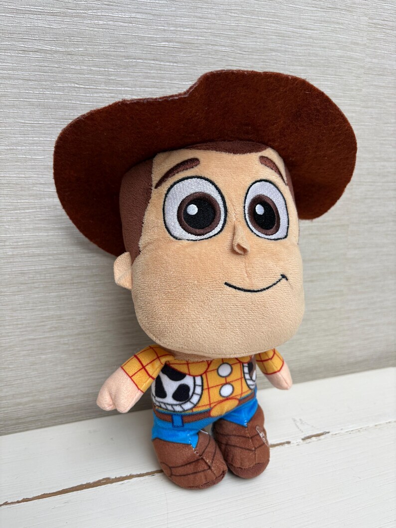 May include: A plush toy of a cartoon character with a brown cowboy hat. The toy has a tan face, brown hair, and large black eyes. It wears a yellow plaid shirt, blue pants, and brown boots. The character has a friendly smile.