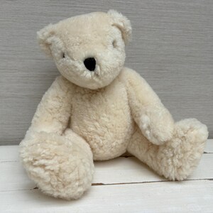 May include: A cream-colored teddy bear with a textured, curly plush exterior. The stuffed animal features a rounded head, small ears, and a dark, solid black nose. It is depicted in a seated position with its arms and legs extended forward.