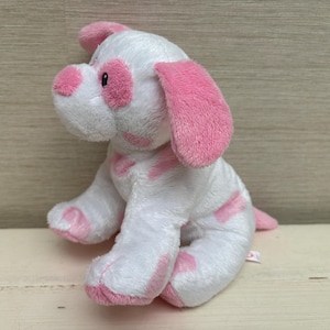 Puede incluir: Un perro de peluche blanco con manchas, orejas y patas rosas. El peluche está sentado sobre una superficie de color claro. El perro tiene una pequeña cola rosa.
