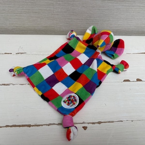 Puede incluir: Una manta de seguridad para bebé suave y cuadrada con un vibrante patrón de cuadros multicolor en rojo, azul, amarillo, verde, negro y blanco. La manta incluye una cabeza de elefante con orejas grandes, esquinas anudadas y un parche circular.